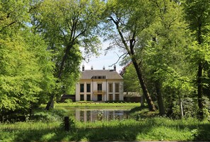 Ein großes, zweistöckiges Haus mit symmetrischer Fassade, umgeben von üppiger Vegetation und einem Teich.