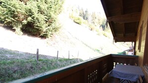 A wooden balcony with a table and chairs, overlooking a grassy area with trees and a road. - Apartment in Wagrain at the ski Lift (Wagrain)
