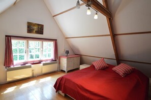 A bedroom with a sloped ceiling, a bed with red bedding, a window with curtains, a radiator, and a small table with a lamp. - Cozy Holiday Home near Forest in Zuidwolde (Zuidwolde)