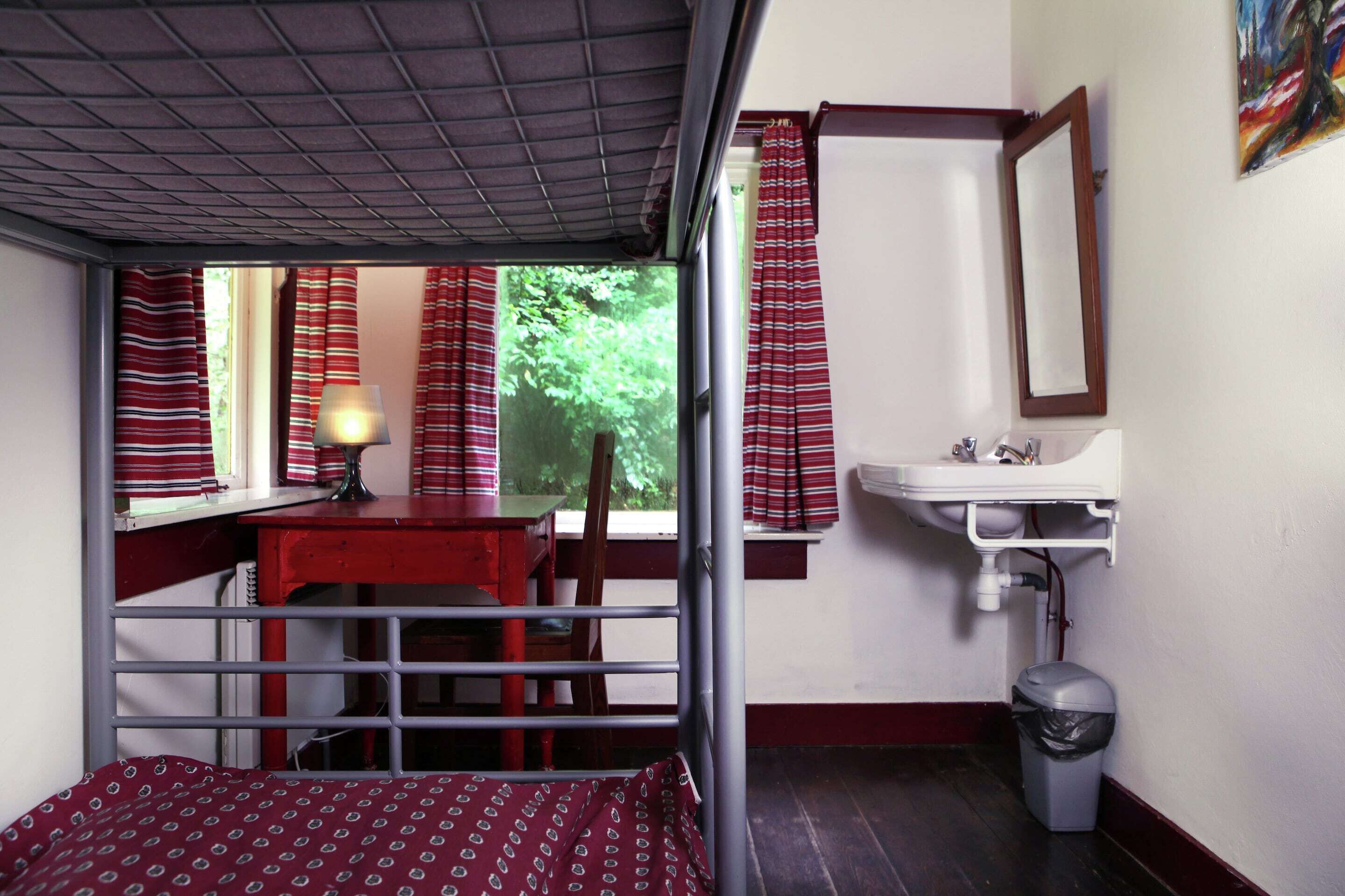 A compact room with a bunk bed, a desk, a chair, a window with curtains, a sink, and a trash bin.