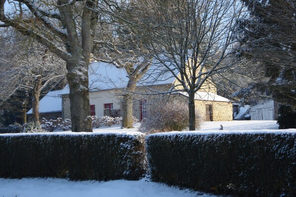 A snow-covered landscape with a stone building, trees, and a hedge.