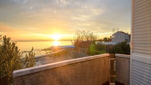 Un balcón con vistas a la puesta de sol sobre una masa de agua, una zona ajardinada y un edificio con tejado inclinado.
