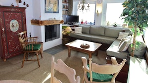 A cozy living room with a fireplace, wooden furniture, a bookshelf, and a large window with a view.