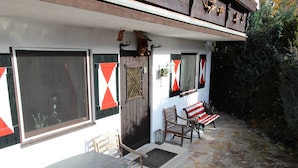 A stone-floored patio with a table and two wooden chairs, a bench, and a door with a red and white triangular decoration.