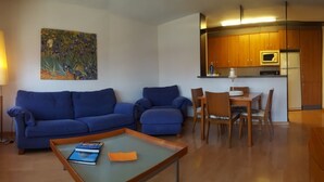 A living room with a blue sofa set, a coffee table with books, a painting on the wall, and a kitchen area with wooden cabinets and appliances. - Duplex 3-3 La Farola (Manresa)