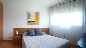 A bedroom with a bed, a nightstand, a lamp, a painting on the wall, and a window with sheer curtains. - Duplex 3-3 La Farola (Manresa)