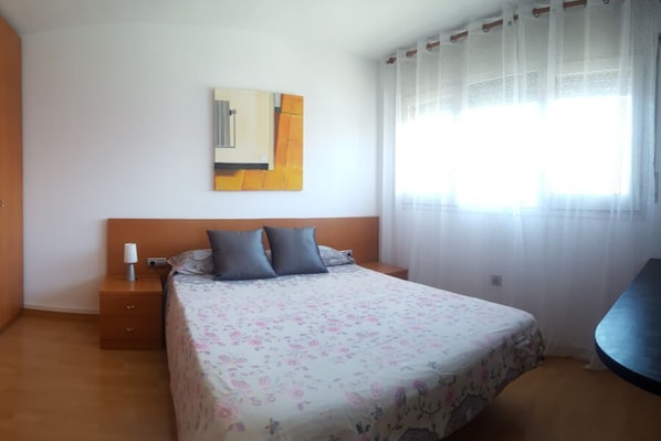 A bedroom with a bed, a nightstand, a lamp, a painting on the wall, and a window with sheer curtains.