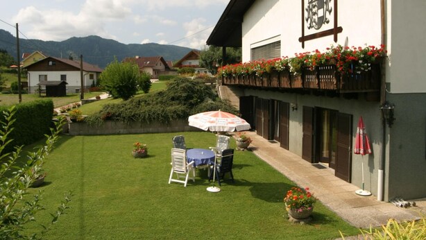 Un edificio con un balcone decorato da fiori rossi, un prato con tavolo e sedie, e montagne sullo sfondo.