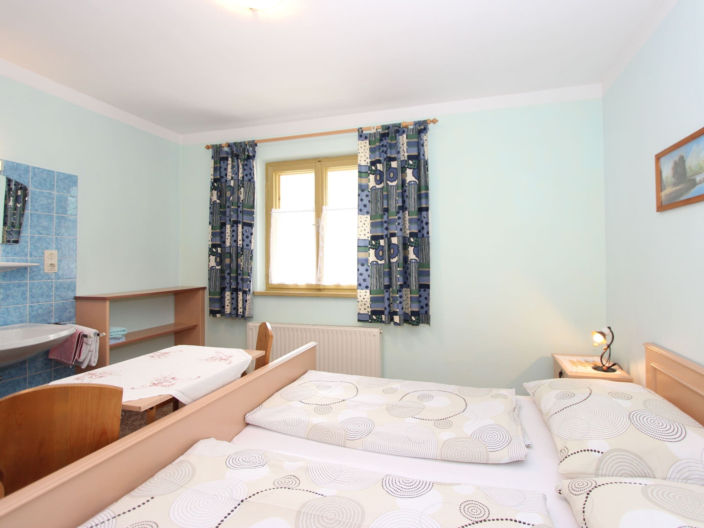 A bedroom with two beds, a small table, a chair, a window with curtains, a radiator, and a wall-mounted shelf.