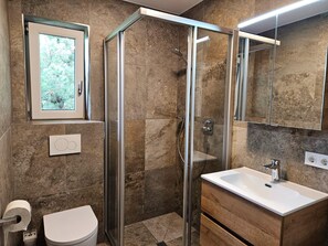 A modern bathroom with a glass-enclosed shower, a toilet, a sink with a mirror, and a window with a view of trees. - Cosy Apartment in Neukirchen near Krimmler Waterfalls (Neukirchen am Grossvenediger)