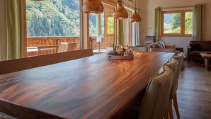 A spacious dining area with a long wooden table, multiple chairs, and pendant lights. Large windows offer views of a forested mountain area.