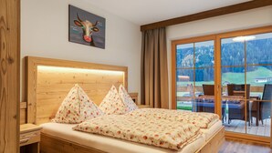 A bedroom with a wooden headboard, a bed with floral bedding, a nightstand, and a view of an outdoor dining area through large glass doors. - Chalet in Wald Near Ski Slopes (Wald im Pinzgau)