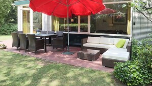 Terrassa exterior amb mobles de vimet, un parasol vermell i una zona de dinar amb finestres de vidre.
