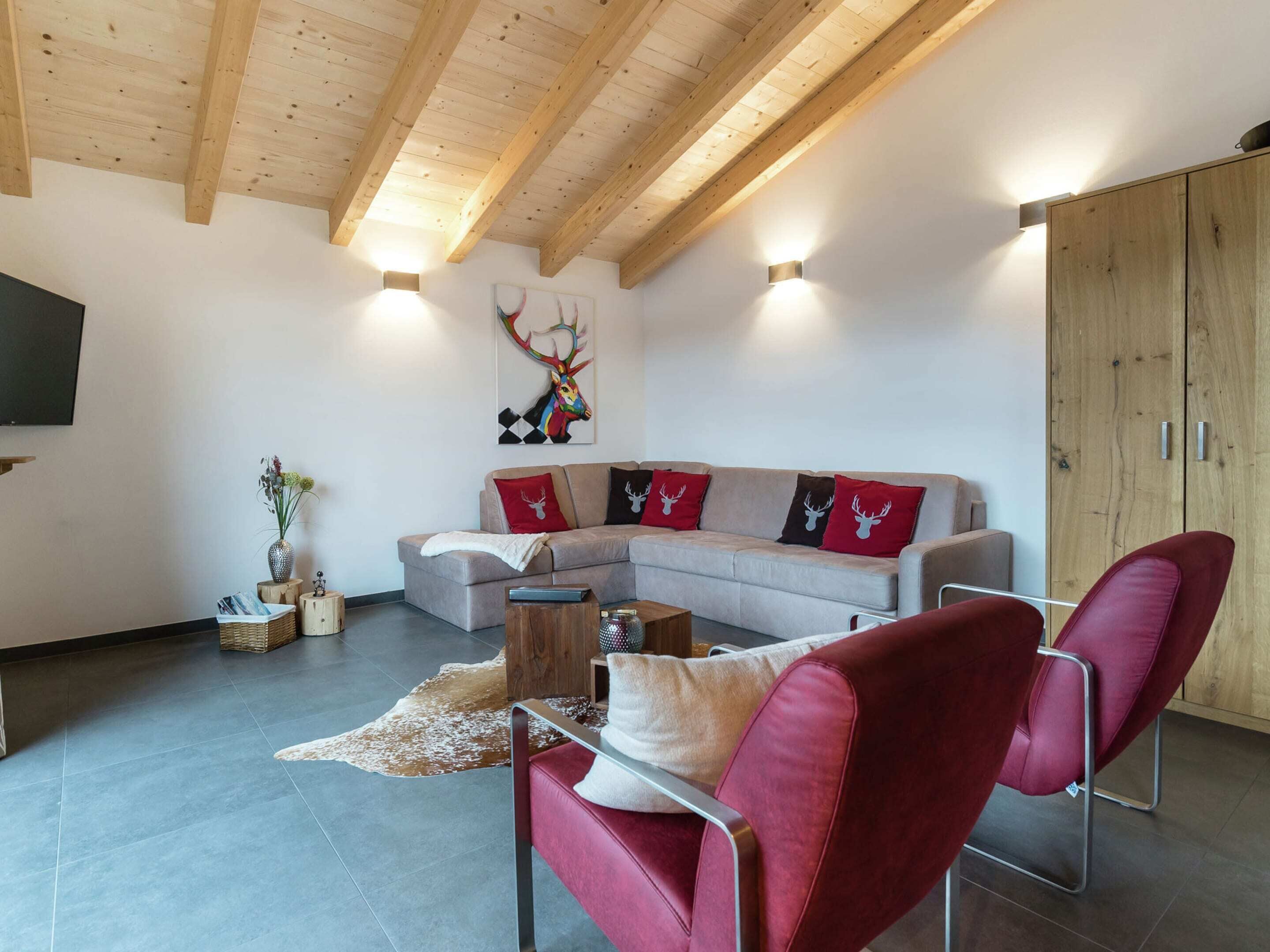 Ein modernes Wohnzimmer mit Holzboden, einem grauen Ecksofa mit Rentierkissen, einem roten Sessel und einem Holzschrank.