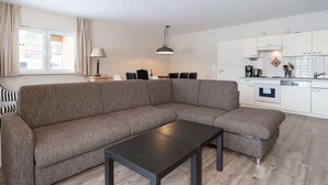 Una sala moderna con un sofá modular, una mesa de centro negra y un comedor con mesa y sillas. Se ve una cocina con gabinetes y electrodomésticos blancos al fondo.