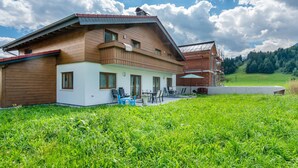Una casa de madera de dos pisos con un balcón, un porche cubierto con mobiliario exterior y vistas a un campo verde y colinas boscosas.