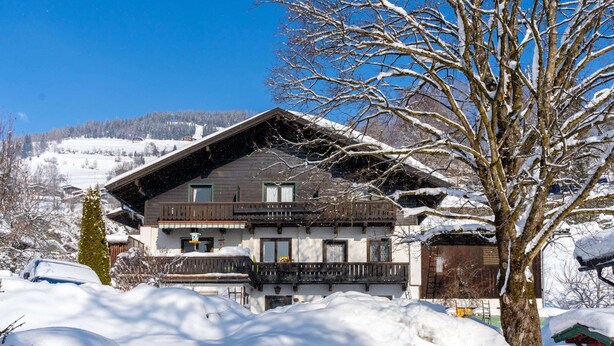 Een chalet met twee verdiepingen en een balkon, omgeven door een besneeid terrein en bomen.