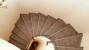 Un escalier en colimaçon aux carreaux gris foncé, doté d’une rampe, mène à un palier où se trouve un luminaire.