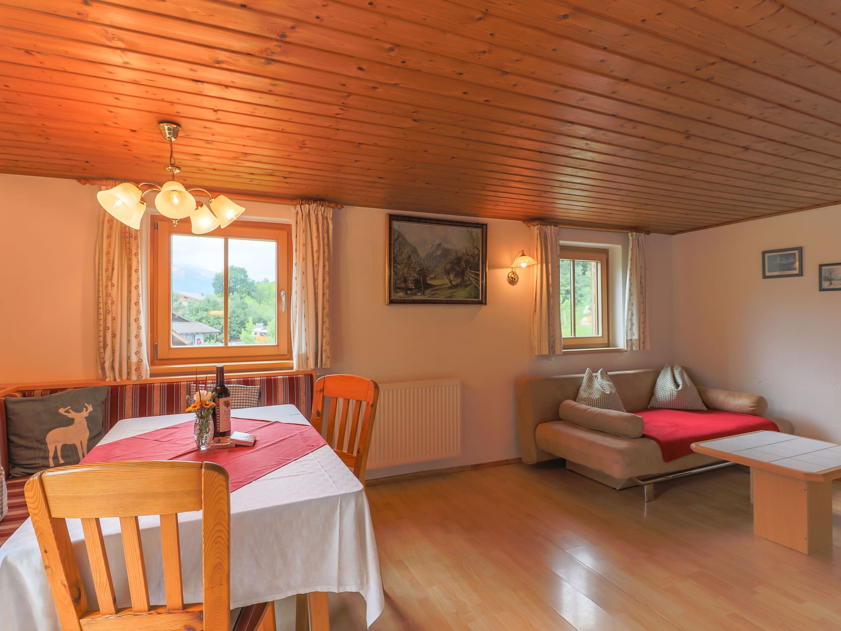 A cozy room with a wooden ceiling, a dining table with chairs, a sofa, and a small table with a red blanket.