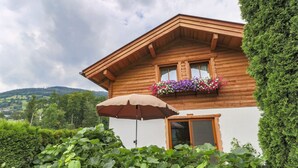 Un chalet en bois doté d’un balcon orné de fleurs aux couleurs vives, d’un grand parasol et d’une végétation luxuriante au premier plan.