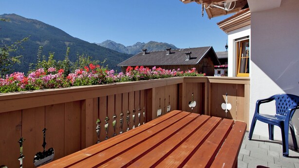 Une terrasse en bois avec un banc et une chaise bleue, d’où l’on peut admirer une montagne et un bâtiment doté d’un toit en pente.