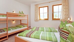 Une chambre avec des lits superposés et un lit simple, un plancher en bois et deux fenêtres avec des rideaux.
