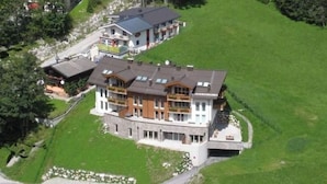 A large, multi-story building with a prominent balcony area, situated on a green hillside with a road nearby.