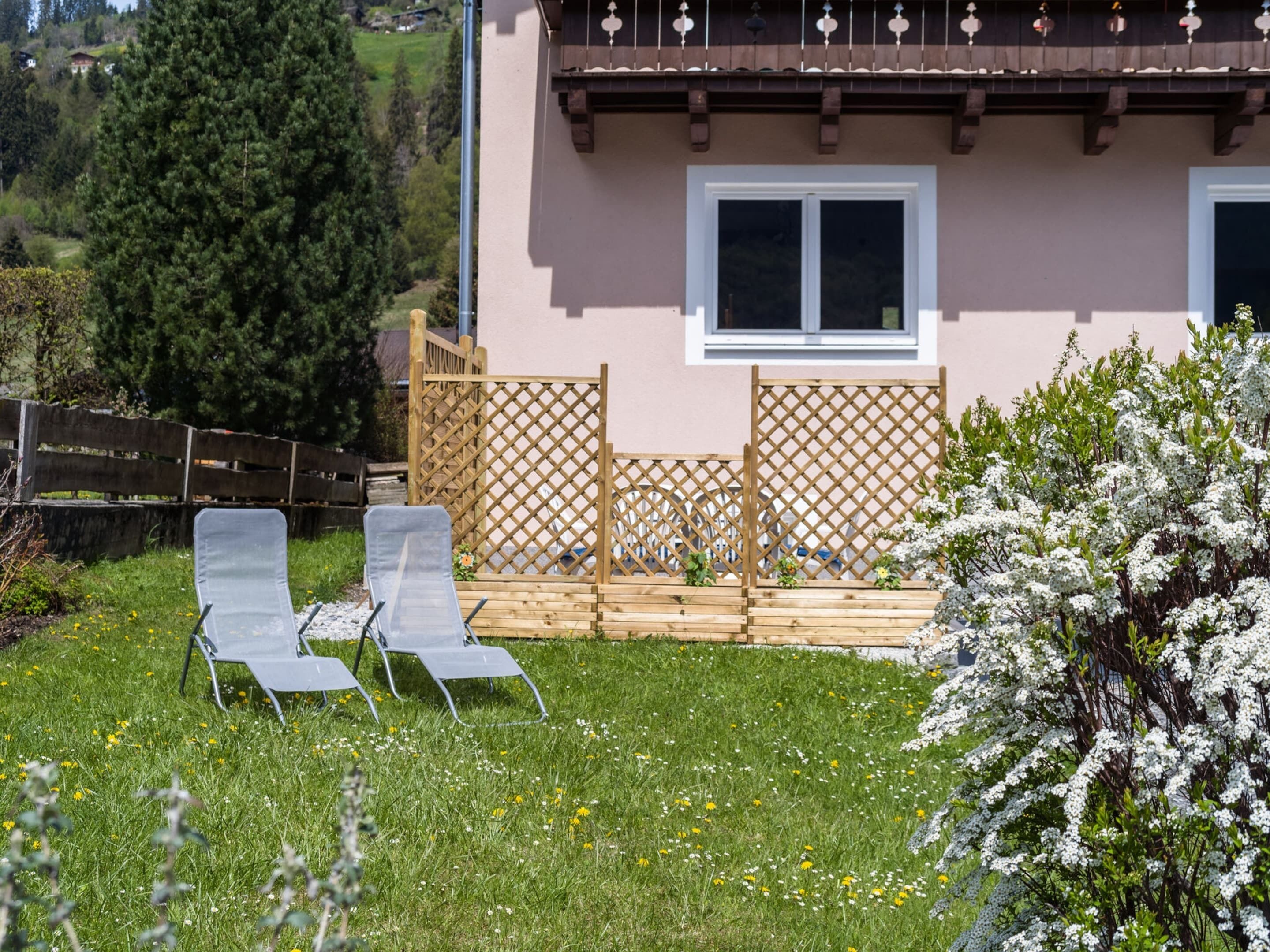 Una casa con terraza de madera, dos sillones de descanso y un jardín con arbustos de flores blancas.