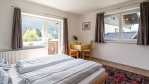 Un dormitorio con cama, dos ventanas con cortinas, una mesita con sillas, un radiador y vistas al exterior.