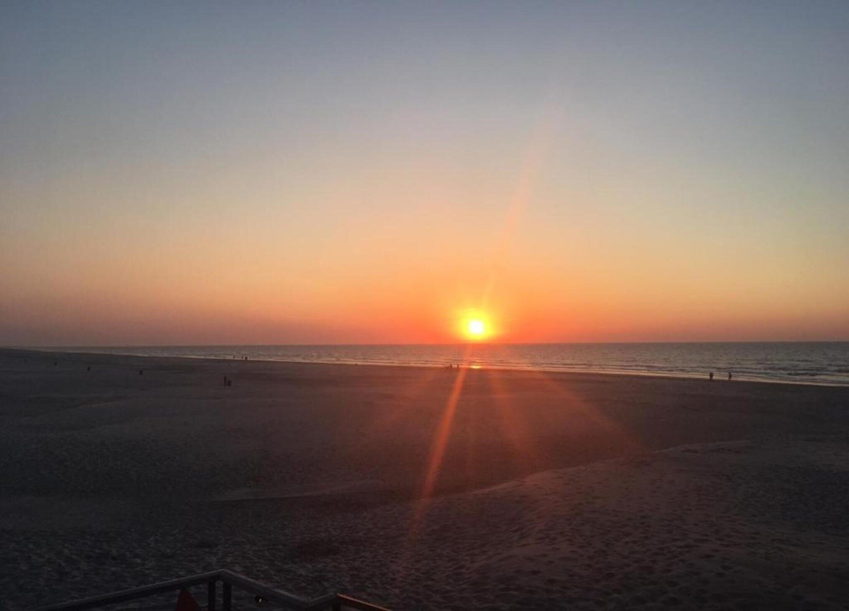 A beach at sunset with the sun low on the horizon, creating a warm glow over the sand and water.