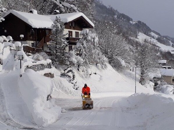 A snow-covered mountain road with a person on a snowmobile. There are wooden buildings with snow-covered roofs in the background.