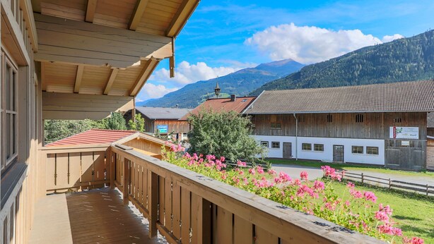 Un balcon en bois offrant une vue sur une montagne, un bâtiment avec un toit rouge et des fleurs roses au premier plan.