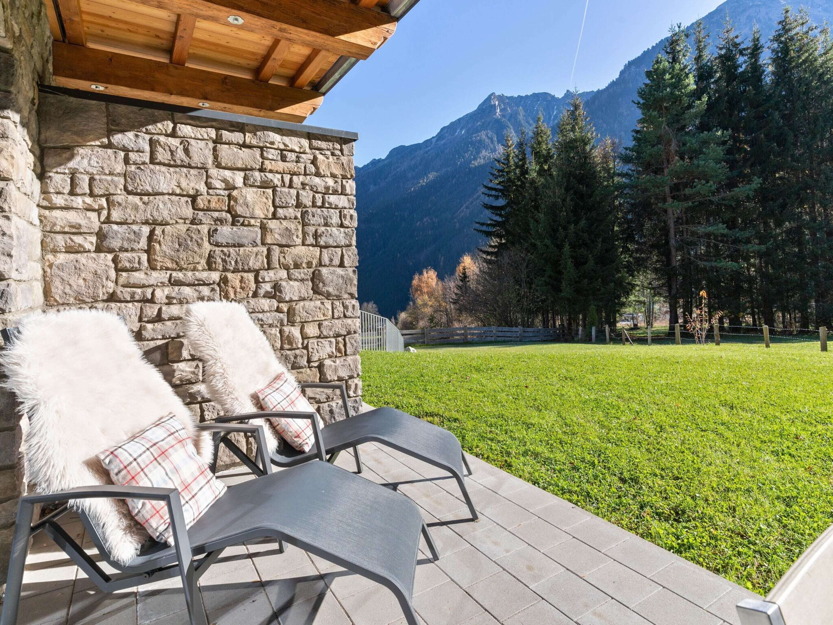 A stone building with a wooden roof, two lounge chairs with white furry cushions, and a view of a mountain range.