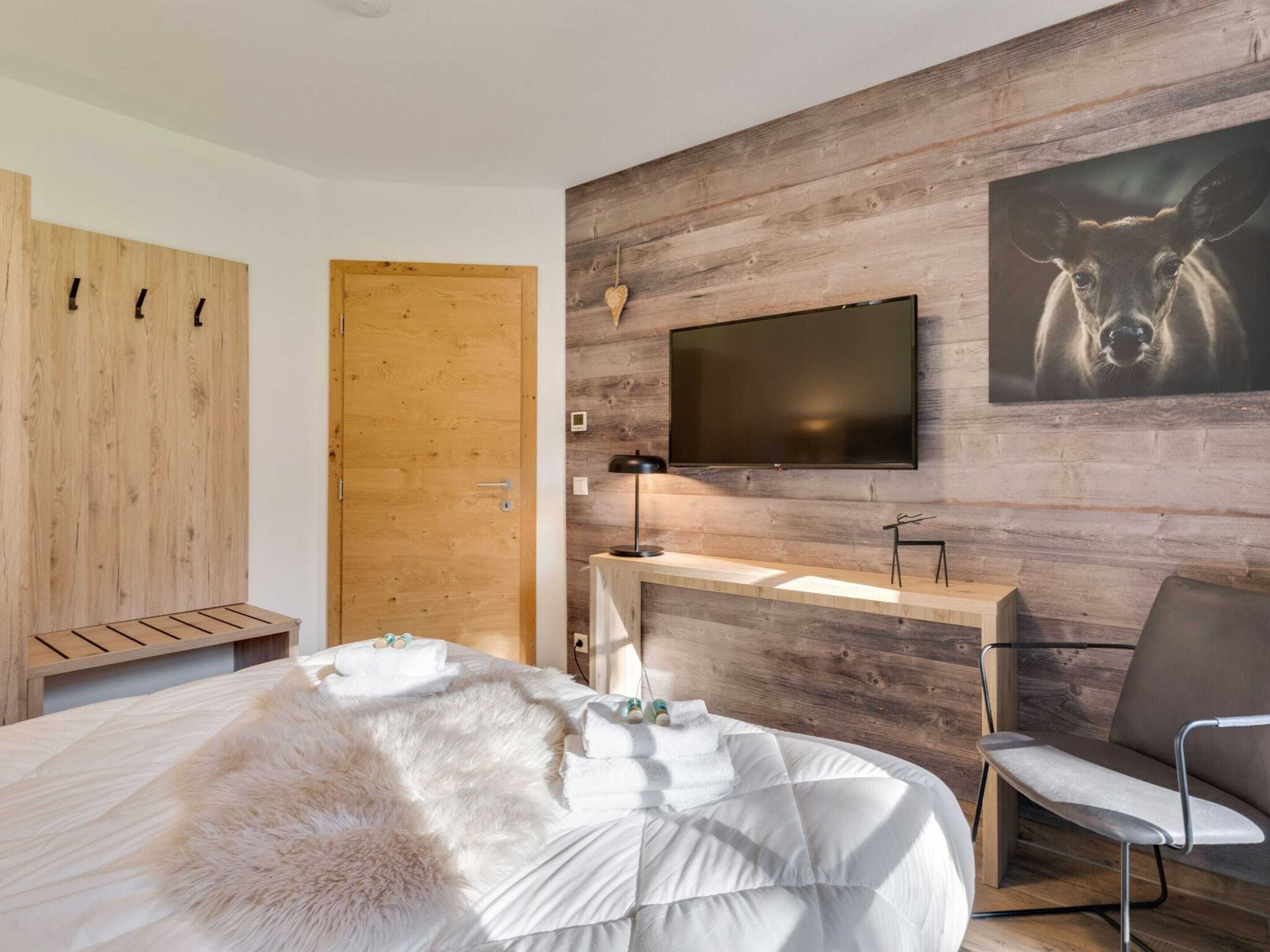 Un dormitorio moderno con pared de madera, un televisor de pantalla plana, cama con ropa blanca, alfombra mullida blanca y una silla.