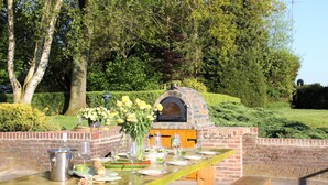 An outdoor dining area with a brick oven, wooden table, and chairs, surrounded by greenery.