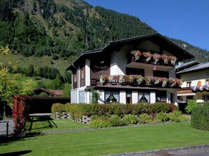 Ein zweistöckiges Gebäude mit einem Blumen geschmückten Balkon, umgeben von gepflegten Gärten und einer Bergkulisse.