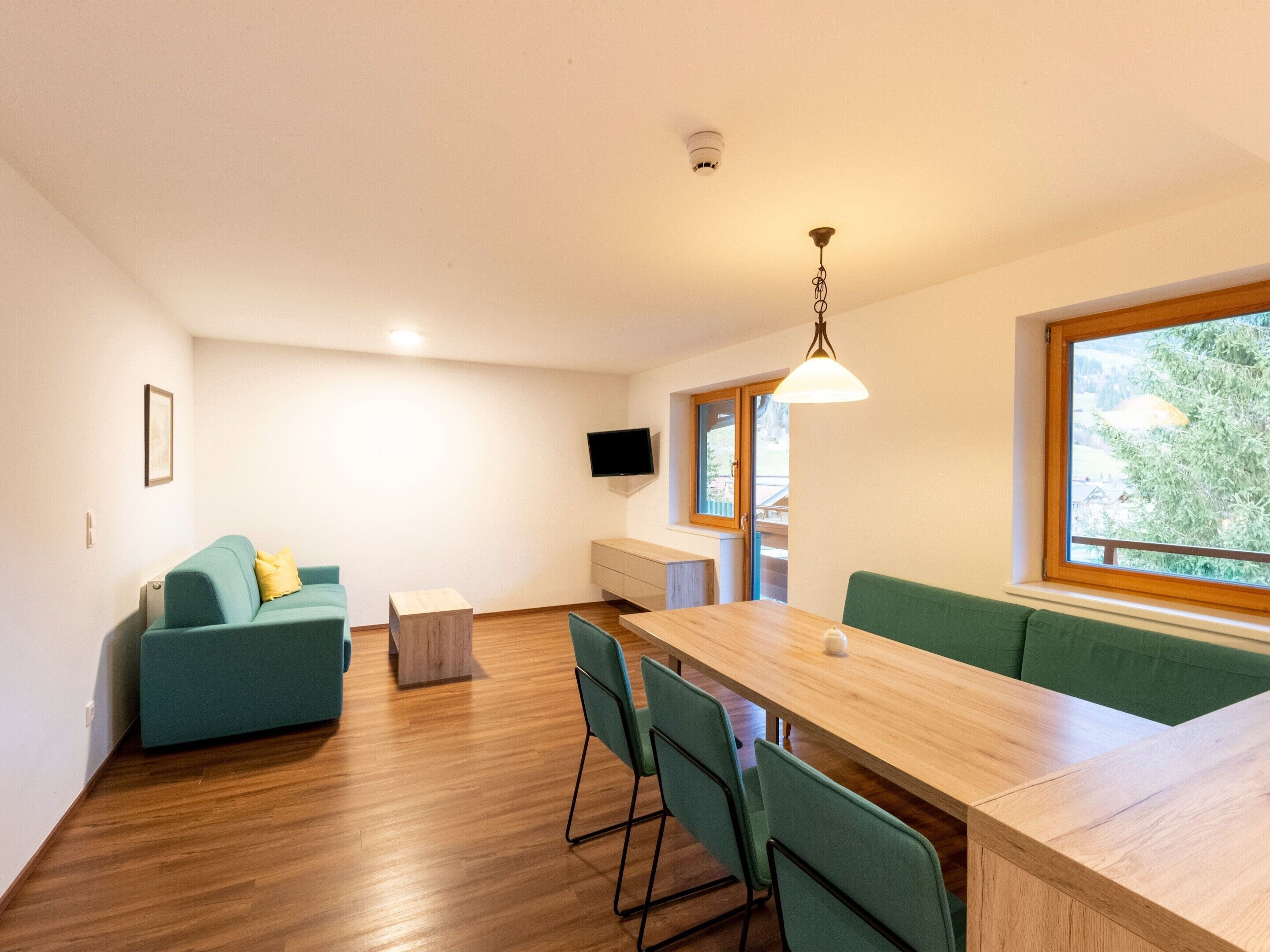 Een moderne, goed verlichte kamer met een houten eettafel, groene bekleding op de zitplaatsen, en een aan de muur bevestigde televisie.