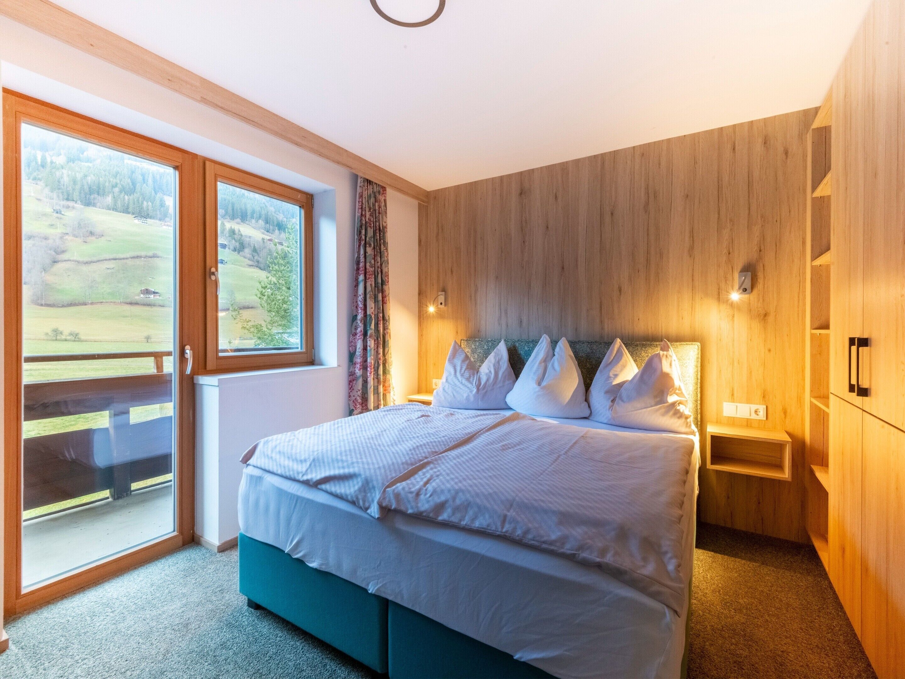 Ein Schlafzimmer mit einem großen Bett, Holzwänden, Blick auf eine grüne Landschaft durch eine Schiebetür und einem kleinen Nachttisch.