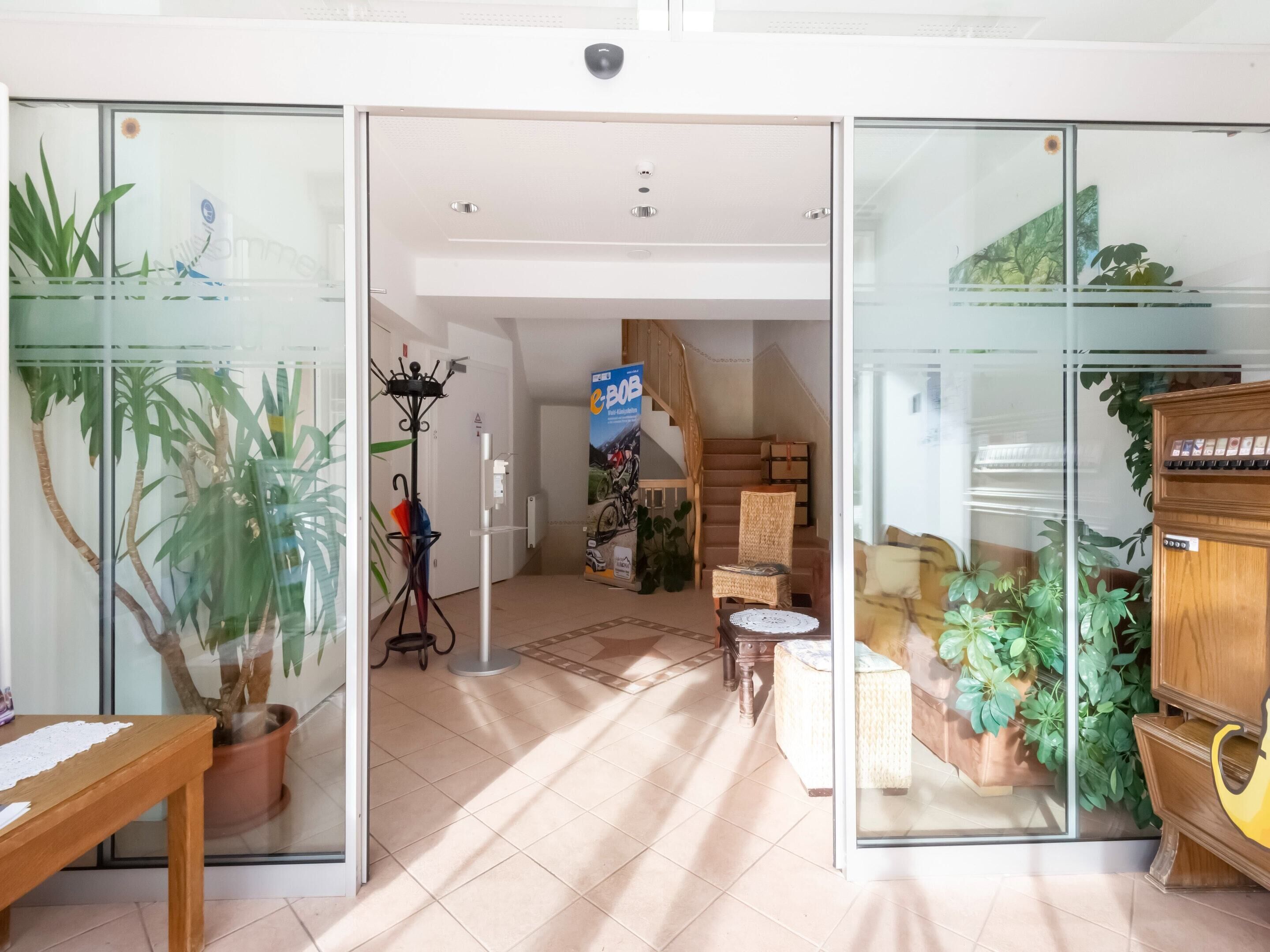 Ein geräumiger Raum mit Fliesenboden, einer hölzernen Bank, einem Blumentopf, einer Glastür, einem Kleiderständer und einer Treppe, die eine weitere Ebene hinaufführt.