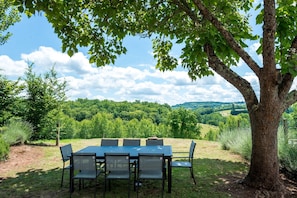 Ein Essbereich im Freien mit Tisch und Stühlen unter einem Baum, umgeben von einer grünen Landschaft und Bäumen.