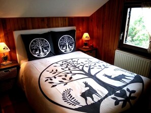 A bedroom with a wooden headboard, a large bed with a tree and deer design, two nightstands with lamps, and a window with a view of greenery. - Spacious Chalet for 4 in Hommert (Hommert)