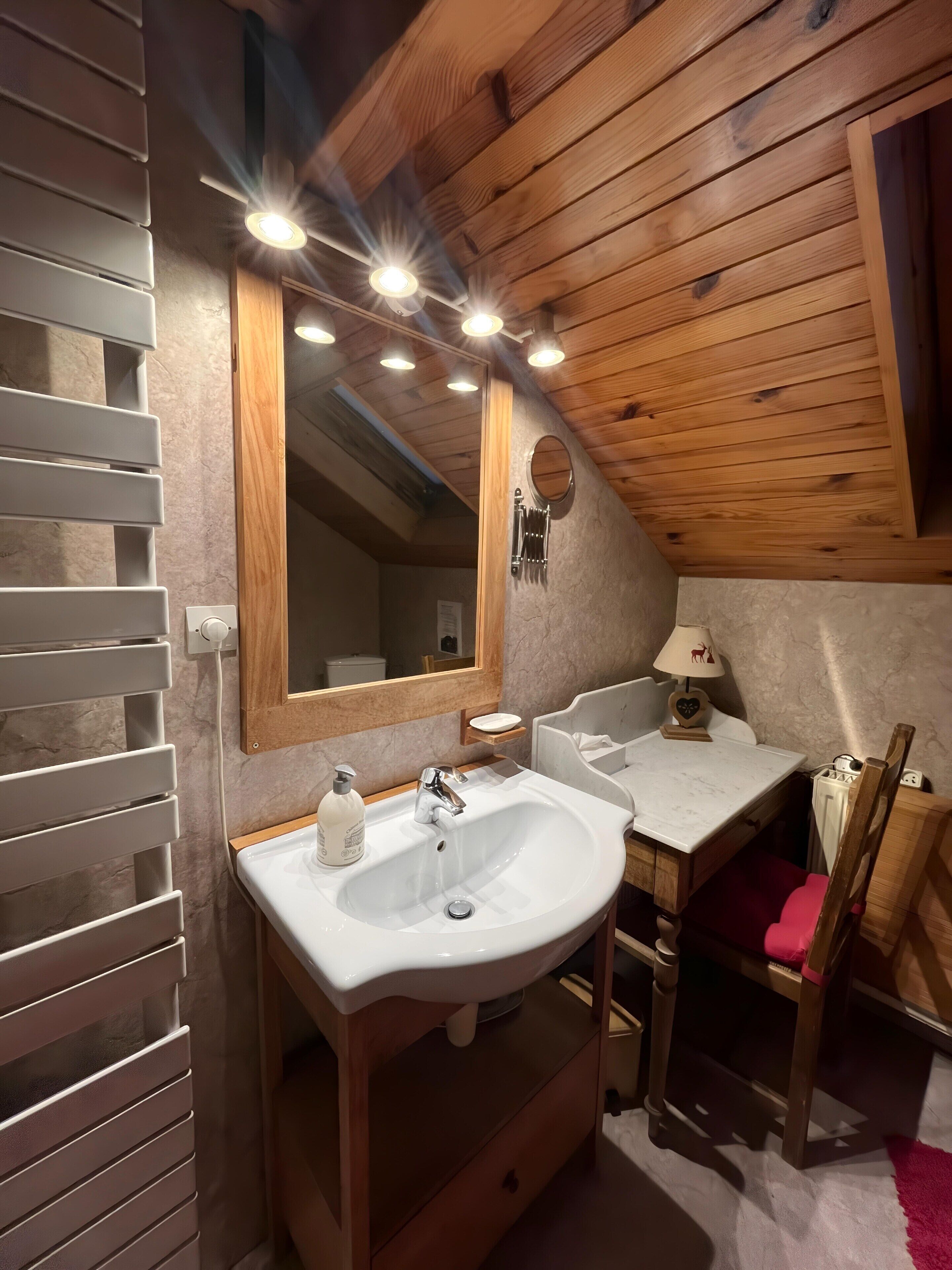 A wooden cabin bathroom with a white sink, mirror, and vanity, a red chair, and a small table with a lamp.