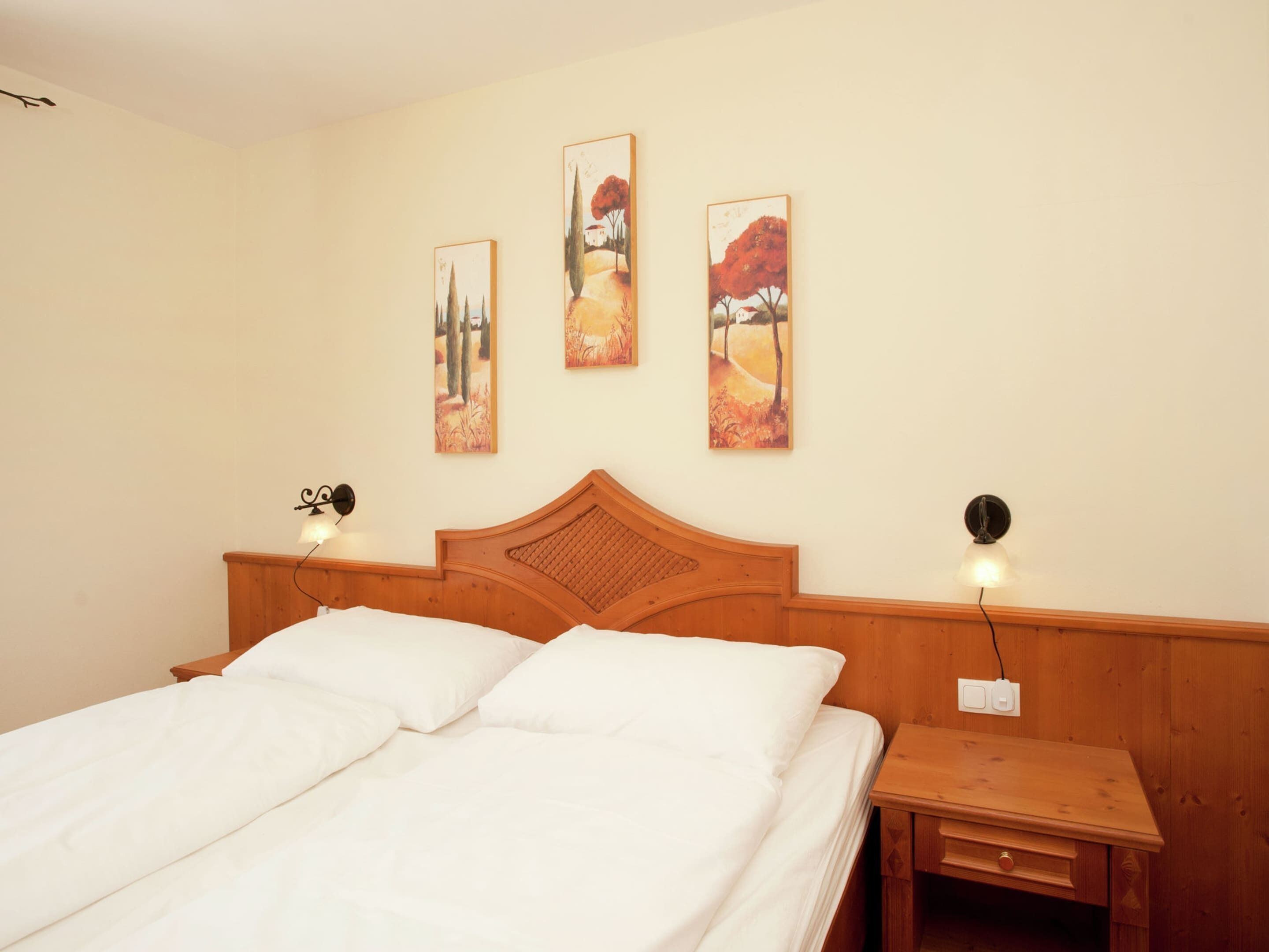A hotel room with a wooden headboard, a bed with white linens, a nightstand with a lamp, and three paintings on the wall.