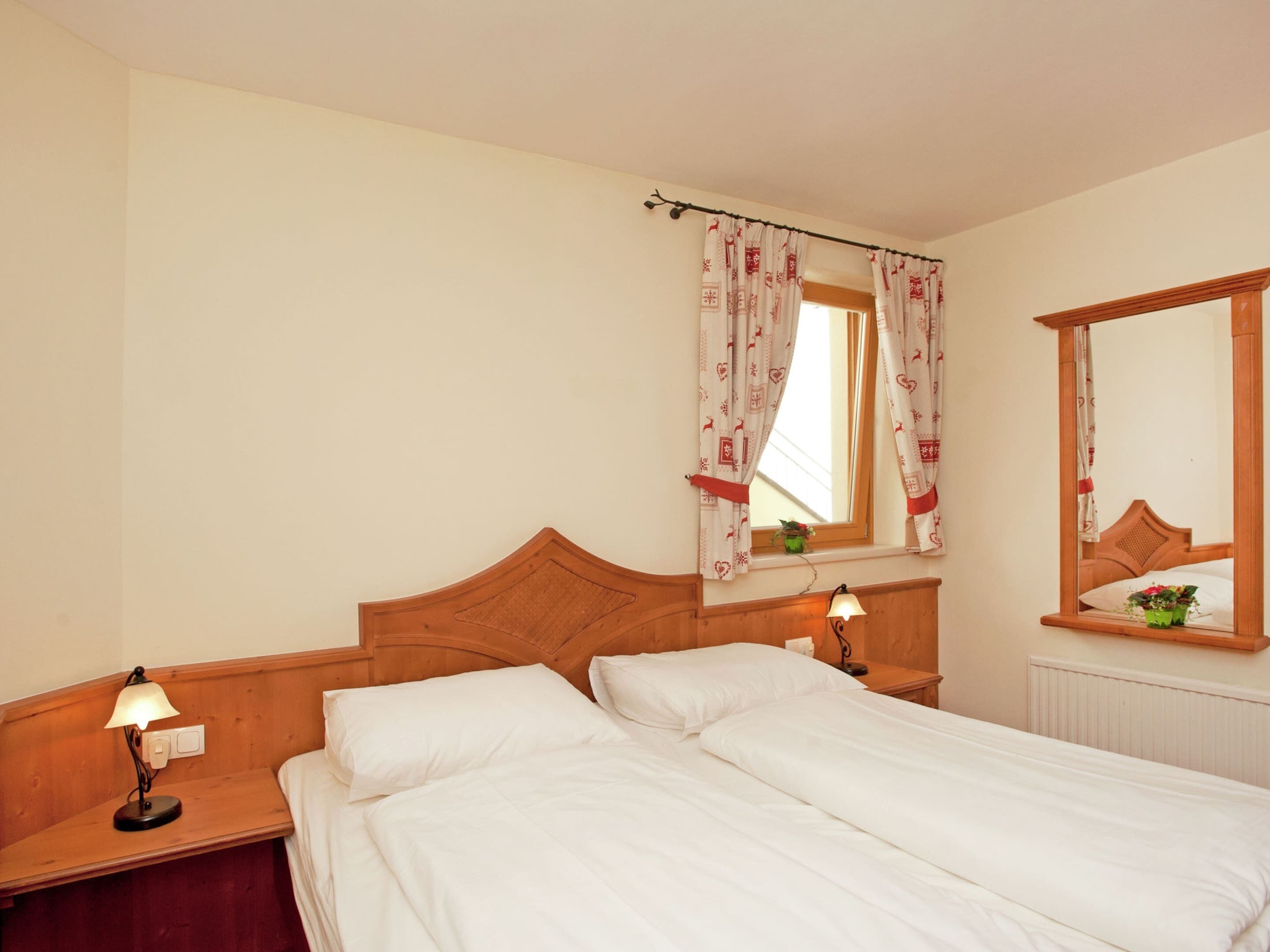 Habitación de hotel con cabecera de madera, cama individual, mesita de noche con lámpara, ventana con cortinas estampadas y espejo en la pared.