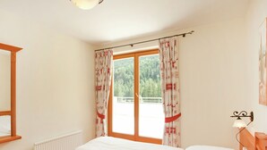 A bedroom with a large bed, a window with curtains, a radiator, a wooden headboard, and a wall-mounted light fixture. - Chalet in Saalbach With Sauna (Saalbach-Hinterglemm)