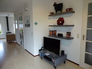 A living room with a TV on a cart, shelves with boxes, a fireplace, and a door. - Holiday Home in Steendam by Schild Lake (Steendam)