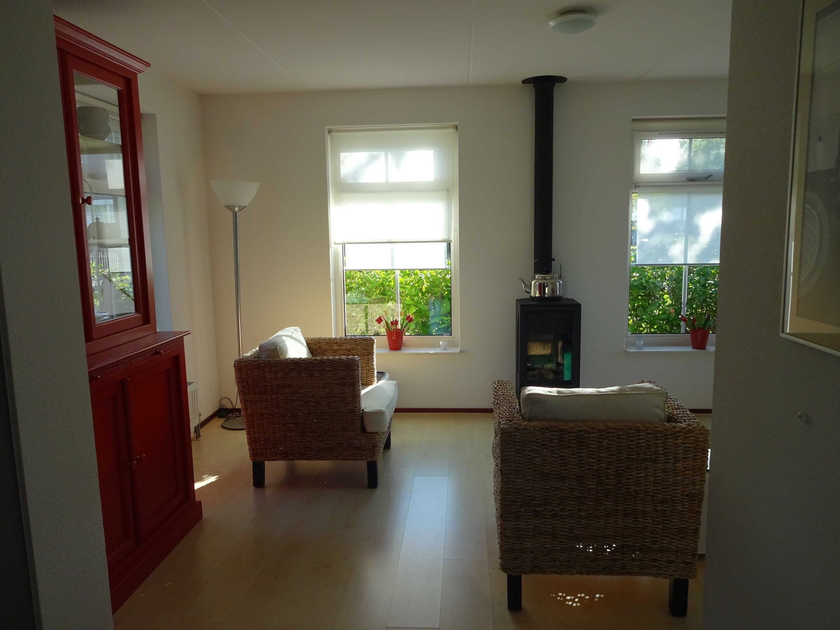 Una habitación con dos sillones de mimbre, un aparador rojo, una lámpara de pie, una chimenea y ventanas con plantas.