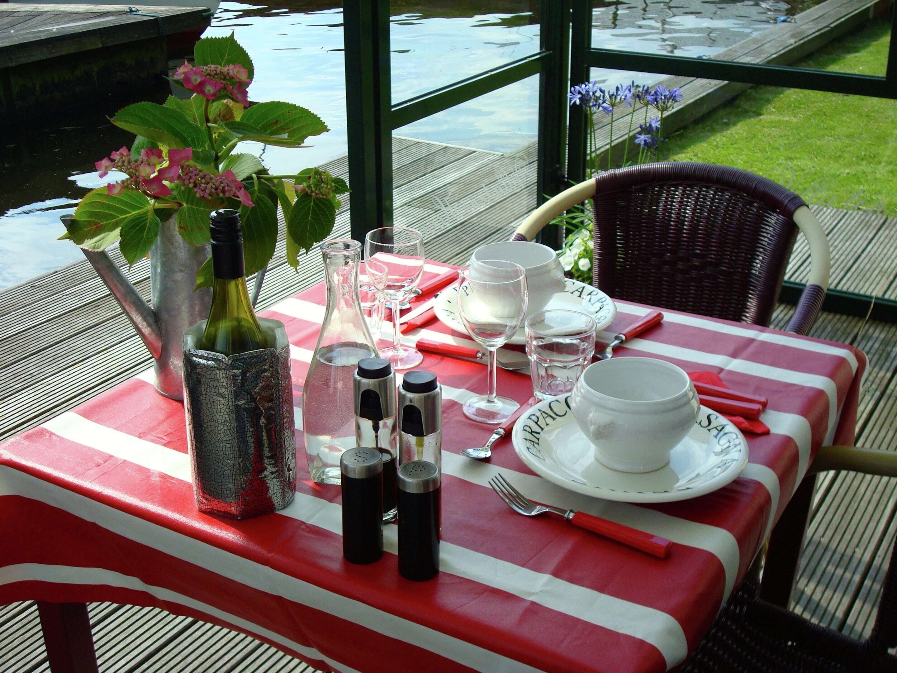 A table set outdoors with a red and white tablecloth, a vase with flowers, wine bottles, glasses, and cutlery.