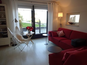Una sala de estar con un sofá rojo, una silla blanca, una mesita redonda con un jarrón de flores y una lámpara.