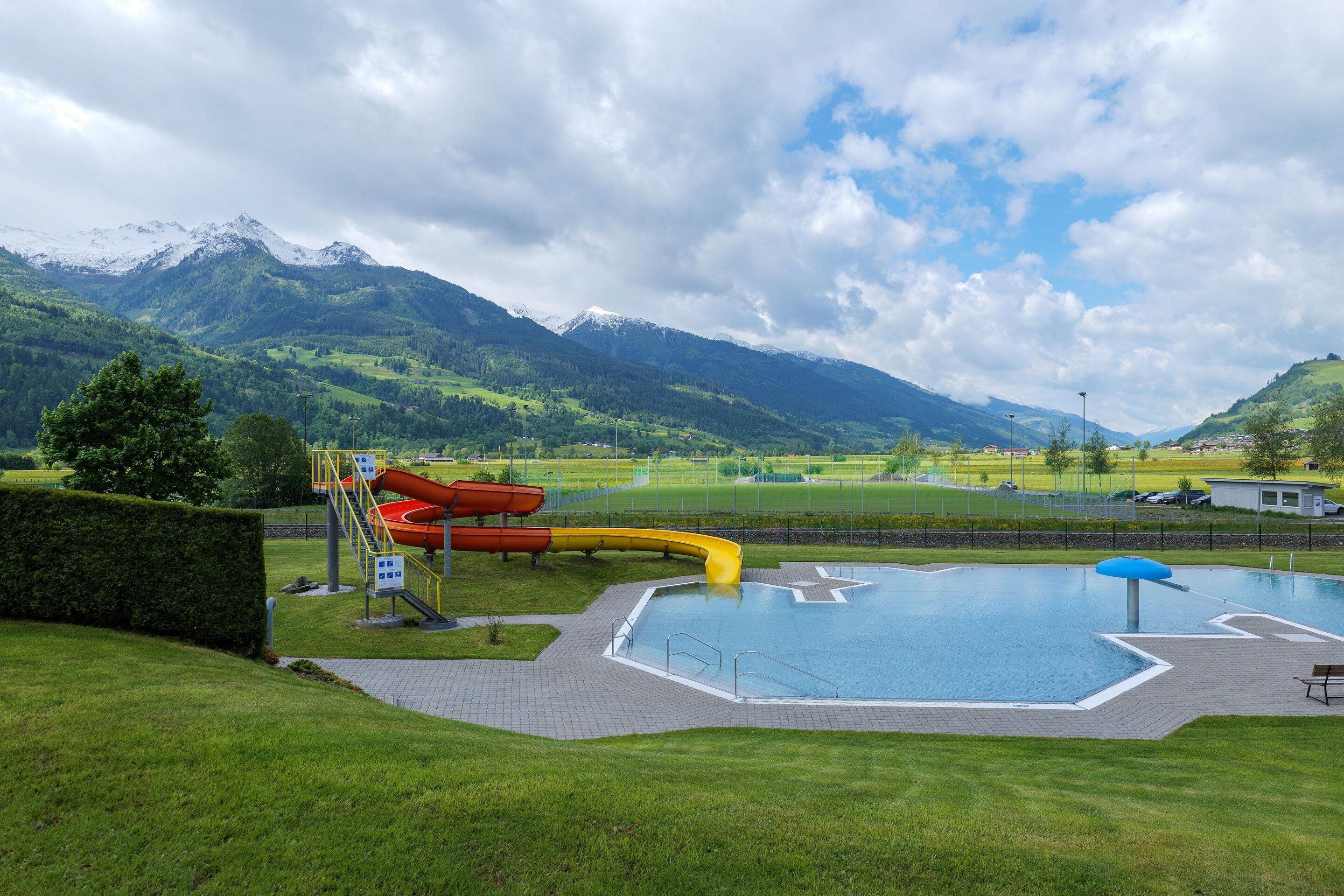 A swimming pool with slides, surrounded by a grassy area and mountains in the background.